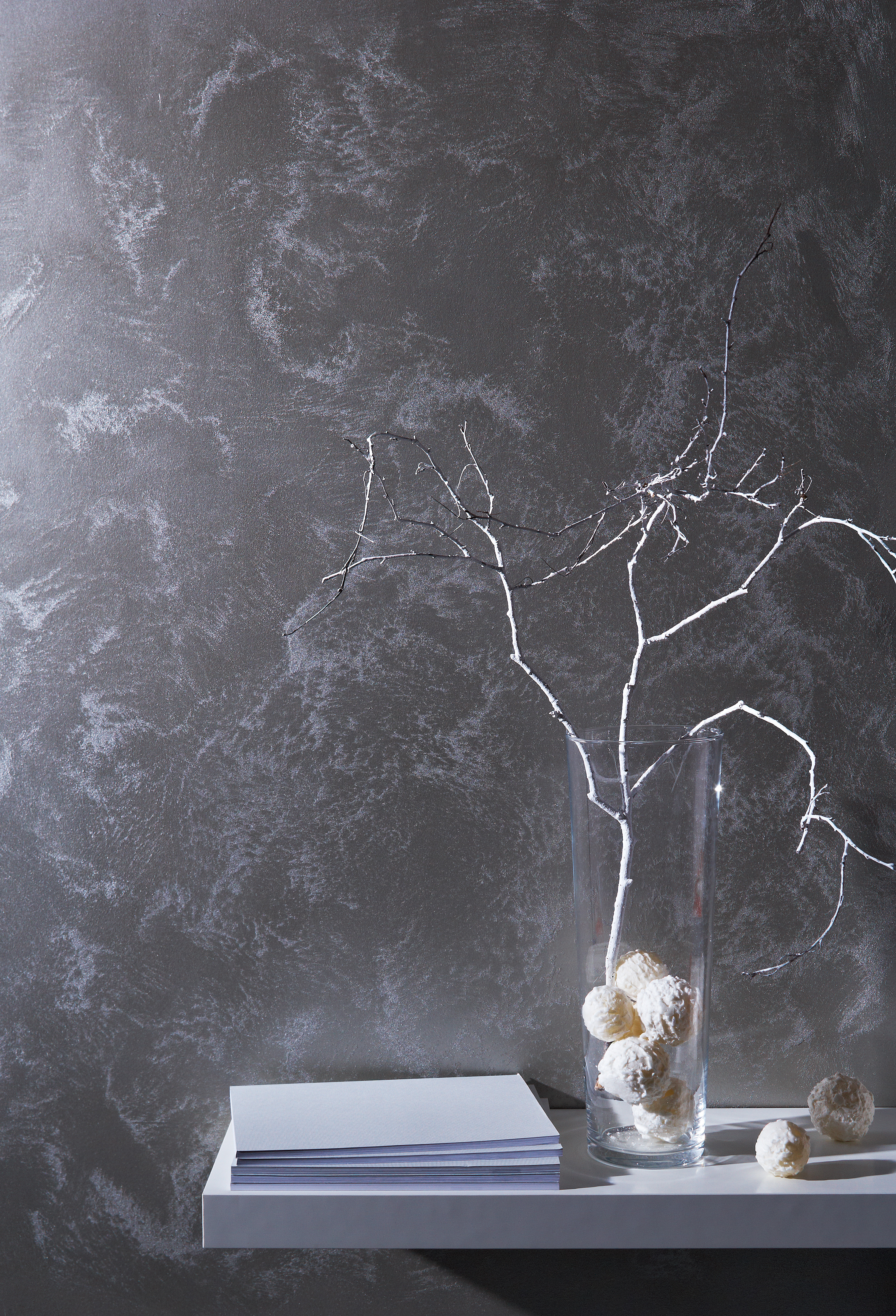 Eine Vase mit einem Zweig und Büchern vor einer gemusterten silbernen Wand